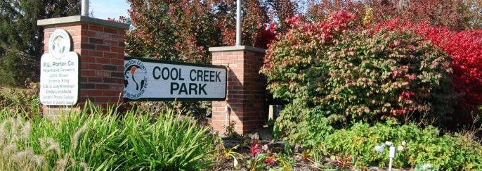 A brick sign reading "Cool Creek Park" stands proudly amidst lush greenery and vibrant red foliage on a sunny day, inviting visitors to enjoy nature's splendor in Hamilton County.